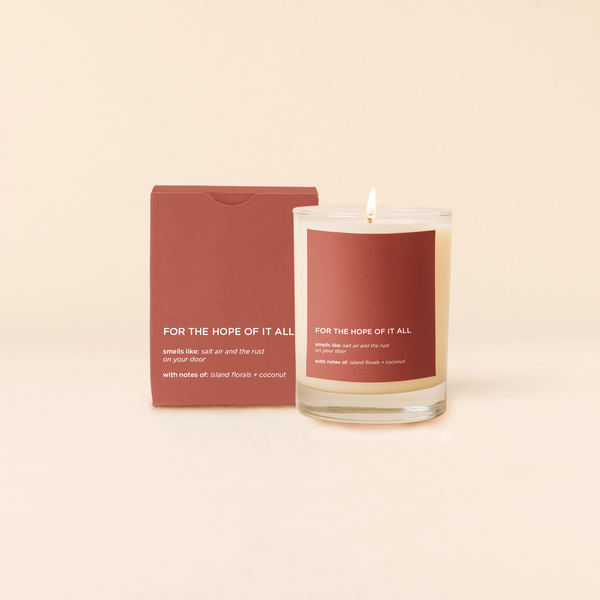 A lit 14oz rocks glass candle with the saying "For The Hope Of It All" written on a brown sticker. There is a matching box in the same brown color with the same saying on it. The candle is all natural soy-based wax (paraffin-free)!! 