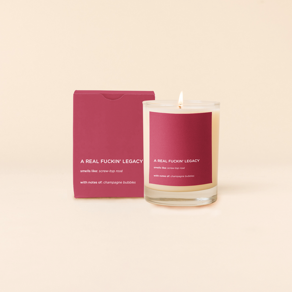 A lit 14oz rocks glass candle with the bold saying A Real Fuckin' Legacy " written on a red sticker. There is a matching box in the red  color with the same saying on it. The candle is all natural soy-based wax (paraffin-free)!! 