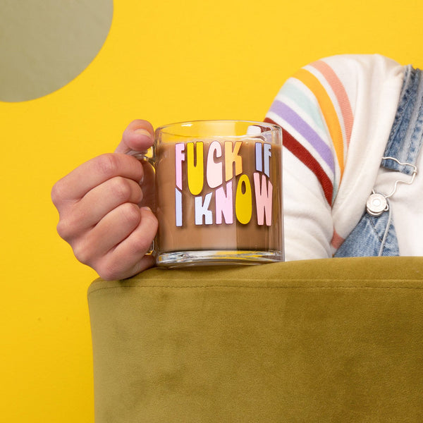 Girl sitting on a couch holding a "fuck if i know" glass mug. Mustard yellow background.