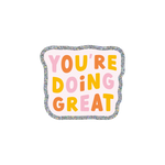 This sticker has a cheerful and encouraging message. "YOU'RE DOING GREAT" is written in all caps in alternating colors of pink, coral, and yellow. The sticker itself has a wavy, cloud-like outline with a silver glitter boarder and a pale pink background.
