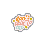 This sassy and playful sticker features a scalloped design with a silver glitter boarder. The text reads "don't talk to me" in orange, pink and light blue colors, written in cursive. There are three olive green smiley faces surrounding the text.