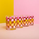 Funky checkered pattern glass cup set in pinks, yellow, and yellow with a liquid inside on a green, pink, and beige background.