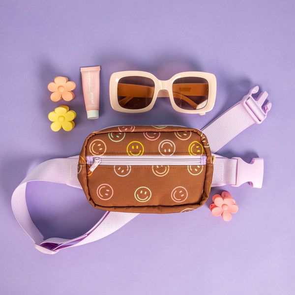 Small brown, colorful smiley faces pattern with a lilac strap hip bag laying on a purple background with misc items 
