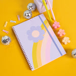 Rainbow flower notebook on a mustard yellow background with small disco balls, jotter pens and misc items.
