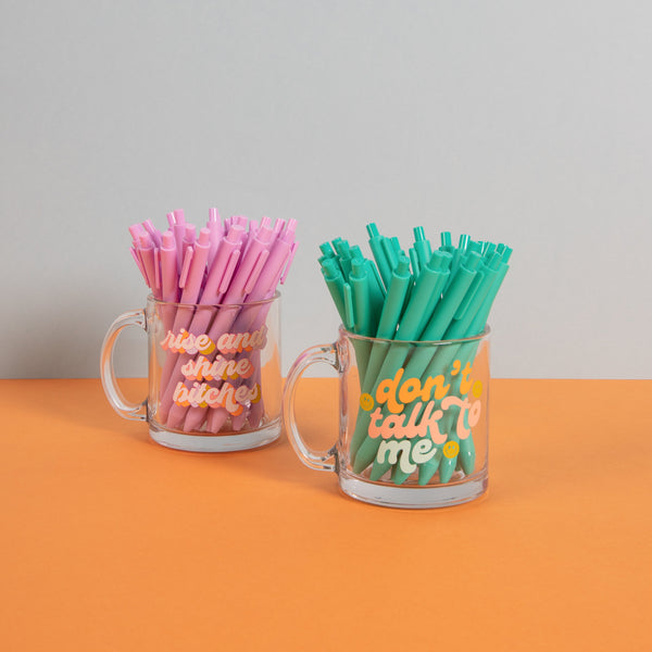 Colorful "dont talk to me" glass mug and colorful "rise and shine bitches" glass mug with jotter pens inside on an orange surface and light blue background