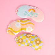 A flatly of 3 weighted eye masks on a blush pink background in an assortment of patterns ranging from wavy checkered boards in citron and periwinkle with daisies scattered around to an ombre back ground with white daisies scattered around, and another that says "No More Mondays" with a rainbow in pink, orange, and yellow above it, a true self care scene.