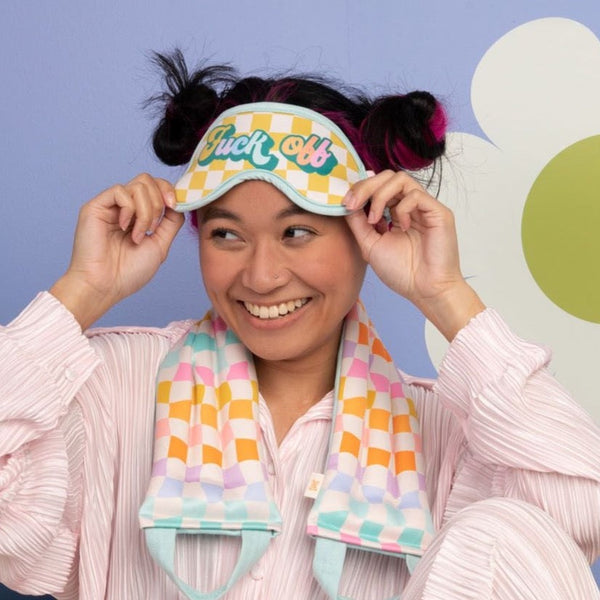 Girl wearing the colorful "fuck off' sleep mask with a colorful carnival checks weighted neck wrap and a purple background with a white flower
