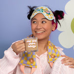 Girl holding "rise and shine bitches" glass mug wearing "fuck off" eye mask and wavy daisy weighted neck wrap. Periwinkle background.