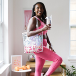 A woman in pink work out gear holding a clear vinyl tote bag filled with multi colored poms. She is holding a clear acrylic tumbler with a straw. Workout gear is shown in the back and a framed poster says "it's ok to start today"