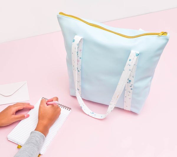 a cute tote bag in light blue vegan leather with white paint splatter straps and zippered top next to a taskpad