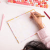 A girl writing with a gold jotter on a large tearaway calendar notepad with a colorful floral border. The notepad is on a white desk alongside a pink and red jotter.