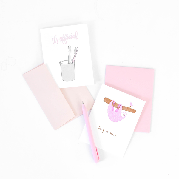 Two greeting cards. The one on the left is a white card with a cup with two toothbrushes and the text "it's official". The one on the right is white with a pink sloth hanging from a branch and the text "hang in there".