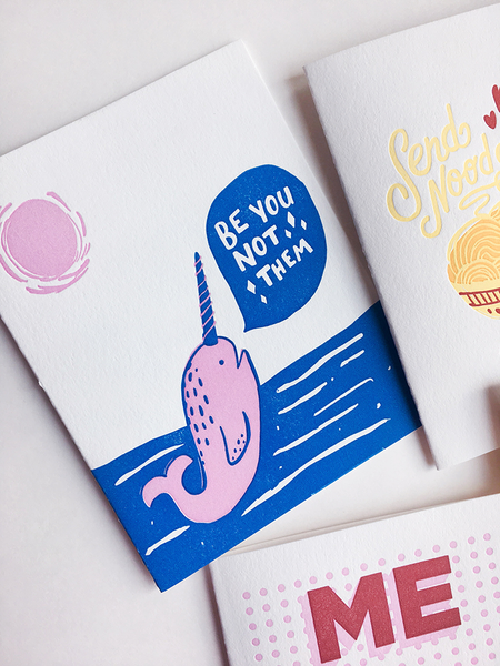 A collection of white greeting cards, partly cropped. The main card has a blue and pink narwhal, blue water and a pink sun. The narwhal has a speech bubble that says "Be You Not Them".