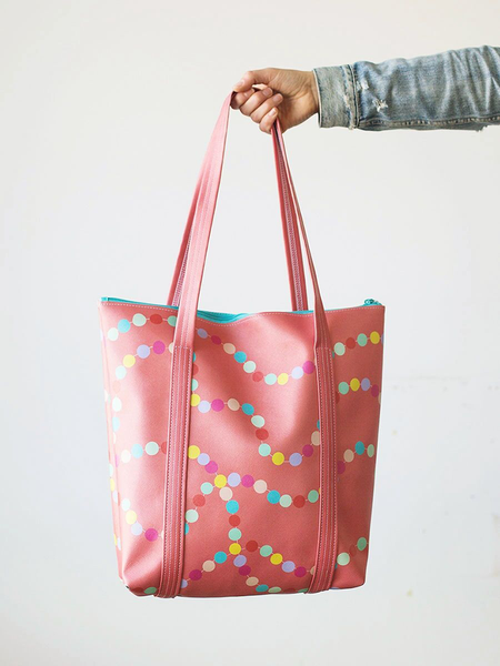 A red tote bag made out of vegan leather. Designed with rainbow dots that are connected and are in curvy lines. Bag is being held by a person in front of a white background.