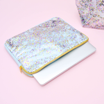 A clear vinyl laptop sleeve with blue canvas and confetti laying on a pink table with a laptop partially out of the laptop sleeve. A second confetti cute pouch is sitting beside the laptop sleeve. 