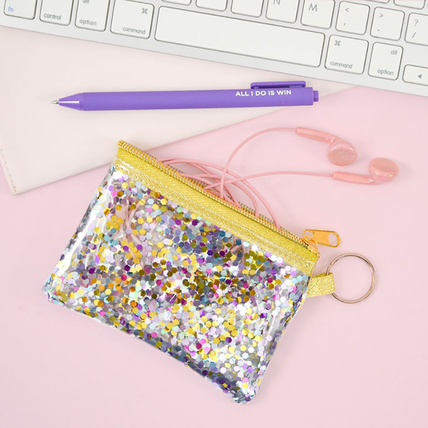 Glitter confetti coin pouch is sitting on a desk with pink earbuds and a purple pen. 