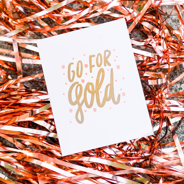 White greeting card with gold text "Go For Gold" and pink stars. The card is laying on metallic ribbon.