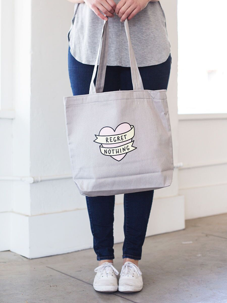Girl in white sneakers holding a cute tote bag in gray canvas with regret nothing banner heart design..