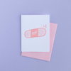 White greeting card with a medium pink and light pink bandaid with the text 