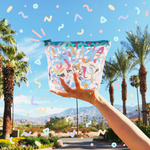 Woman stands holding clear vinyl bag with colorful wiggles drawn on it, with a blue sky and palm trees in the background