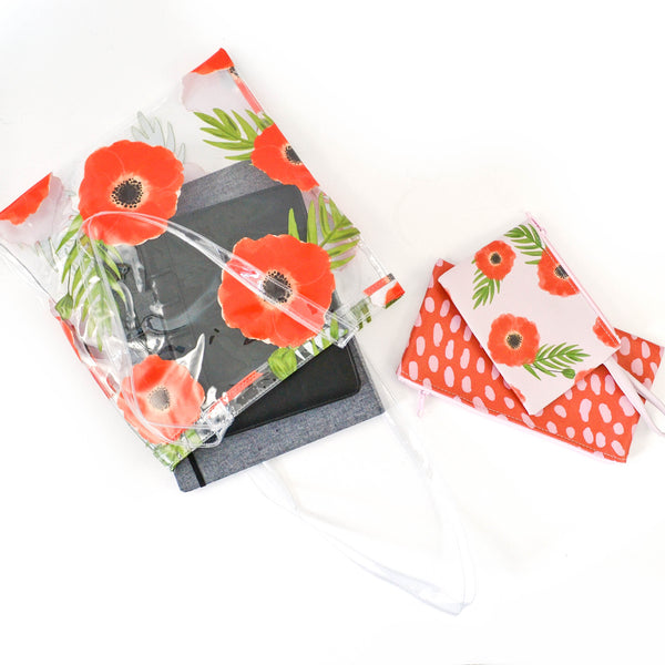 A clear vinyl bag with a red poppy pattern lays on a white surface with a small iPad visible through the bag, with two small pouches sitting beside it. 