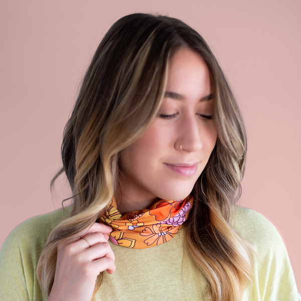 An orange, mask-like item, with multicolored and differently designed flowers that is multifunctional. Displayed by person wearing it around their neck in front of a muted pink background