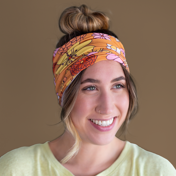 An orange, mask-like item, with multicolored and differently designed flowers that can be used as a headband. Displayed by person using neck-fluff as a headband. 