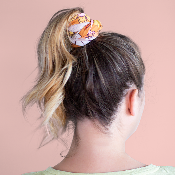 An orange, mask-like item, with multicolored and differently designed flowers being used as a hair-tie. Displayed by person wearing a pony tail and using neck-fluff to keep their hair tied up.