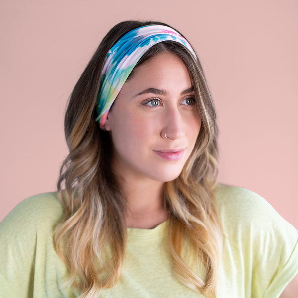A rainbow tie-dye neck/face cover that is being used as a headband on a woman's head in front of a dusty pink background.