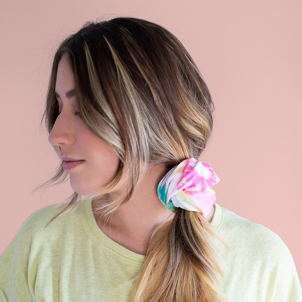 A rainbow tie-dye neck/face cover being used as a hair tie on a woman in front of a dusty pink background.