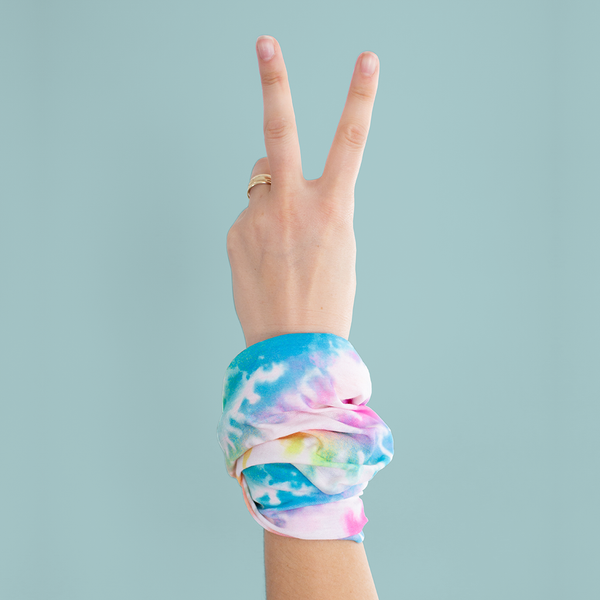 A rainbow tie-dye neck/face cover wrapped around a persons wrist that is in front of a muted blue background.
