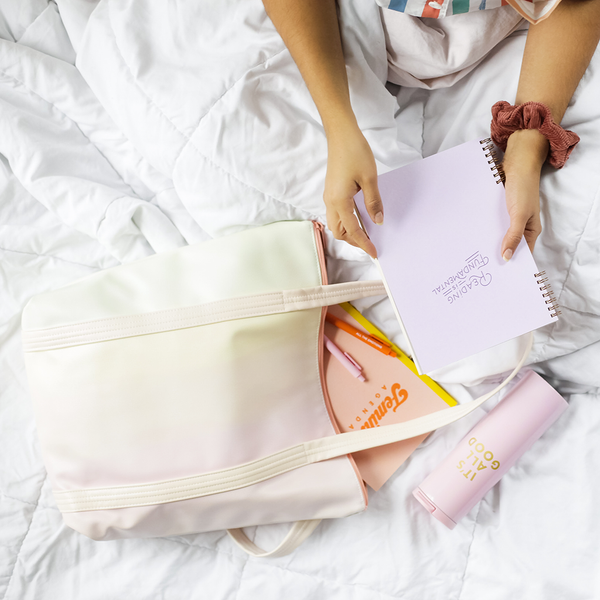 A woman's hands holding a spiral bound notebook with "Reading is fundamental" printed on the cover. There is a pastel gradient vegan leather tote bag with jotter pens and a notebook printed with the text "Feminist Agenda" printed on the cover. There is a light pink stainless steel tumbler printed with "It's all good". 