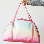 Girl's hand holding a cute travel bag in rainbow ombre with coral straps.