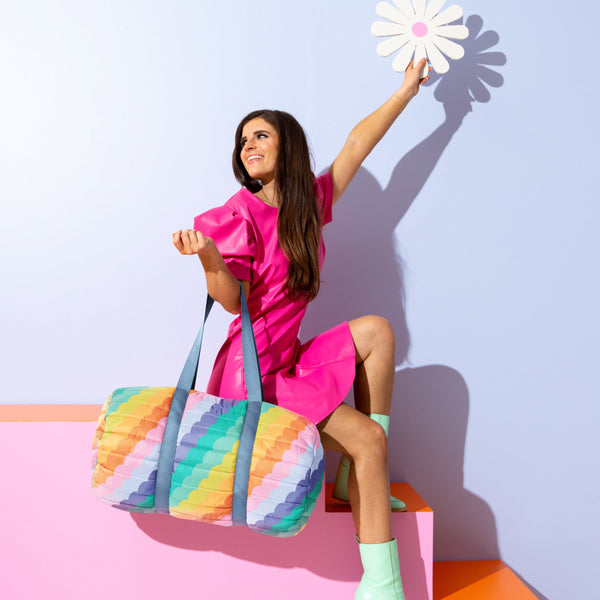 Young lady sitting on a stair case with a rainbow scalloped putt duffle bag on her wrist. 