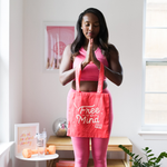 A woman appears to be meditating with her hands together. A coral color canvas tote bag with the text printed "Free Yo Mind" is hanging from her hands. 