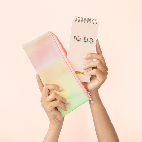 A pastel rainbow ombre pouch with a peach pink zipper. A to-do task pad is being taken out of pouch and both items are displayed in front of a light pink background.