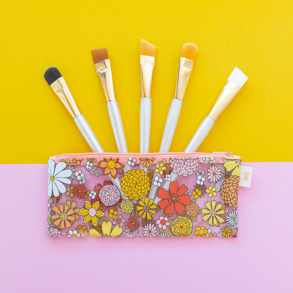 A clear rectangle-shaped pouch with retro-style flowers printed all over. Inside the pouch, there are 5 paintbrushes with blue handles and a gold ferrule, and it is all against an orange and pink background.