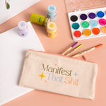 A cream-colored canvas pouch with the phrase "Manifest That Shit" on the front in multi-colored font, the phrase is surrounded by minimalist sparkle stars. 