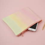This large pencil pouch is a pastel ombre pattern with a pink zipper. Ombre goes from a peach pink to a light green. Displayed in the pouch is a phone along with a gold colored Jotter pen. All items are on a pink background.