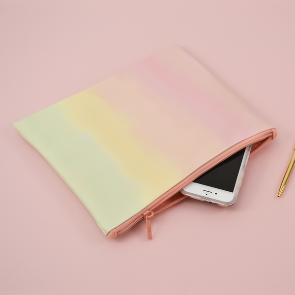 This large pencil pouch is a pastel ombre pattern with a pink zipper. Ombre goes from a peach pink to a light green. Displayed in the pouch is a phone along with a gold colored Jotter pen. All items are on a pink background.