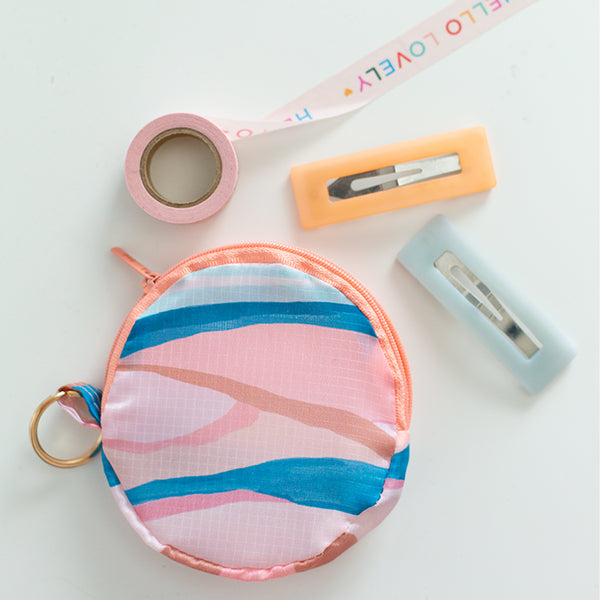 An abstract, wave-like design circle pouch with a zipper and a keyring. The pouch is in pastel and neutral colors. There is also washi-tape and hair clips displayed with the pouch.