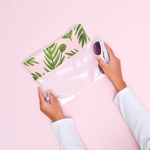 Woman holding a vegan leather and clear vinyl zippered pouch. The vegan leather is printed in pink with greenery all over. One of the woman's hands is holding onto white sunglasses. 