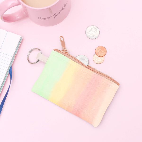 A rainbow ombre pouch with a peach zipper and a silver key ring. Displayed with a mug and some coins in front of a pink background.