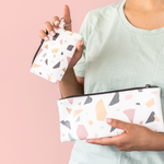 A white small pouch with terrazzo specs printed all over. Small pouch has a keyring and is being held up by a person who is also carrying a larger pouch with the same design.