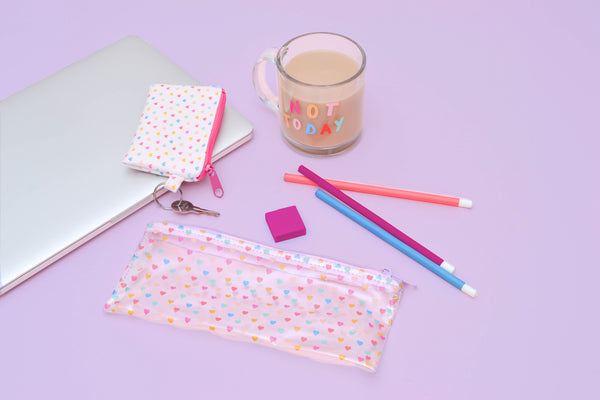 A clear vinyl pouch with tiny heart illustrations sits on a pink surface next to a cup of coffee, a computer, three pencils and a matching smaller pouch attached to a key.