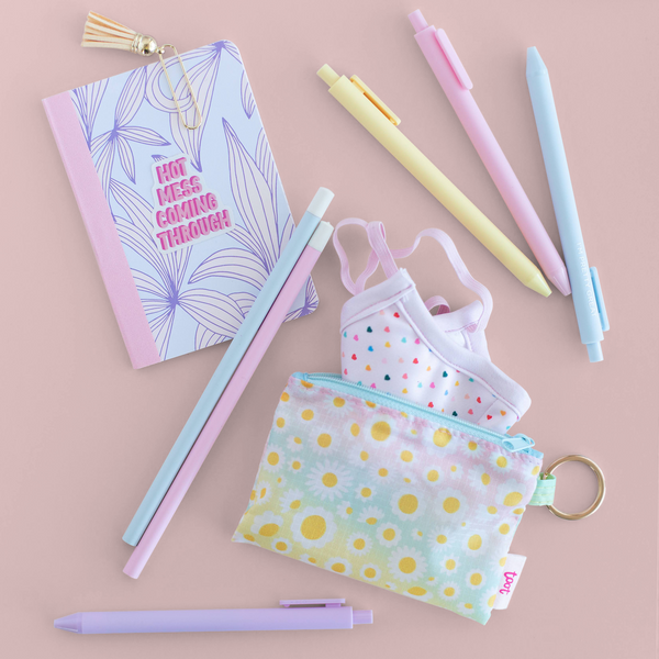 A small ombre pouch with white daisies all over and a facemask with hearts on it next to a mini notebook, pencils, and pens