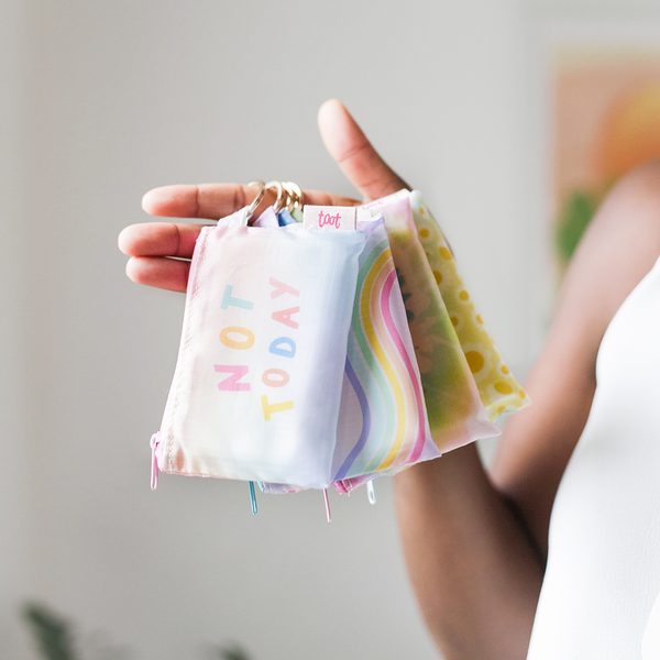 A woman's hand holding four ripstop printed zipper pouches with key rings. The pouch in front reads "Note Today". The second pouch is a wavy rainbow pattern. The third pouch is a tie dye pattern. The fourth pouch is a daisy pattern.The key rings are hanging from her index finger. 