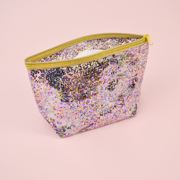Large cosmetics bag in clear vinyl with glitter confetti lining, a gusseted bottom and gold zippered top closure.