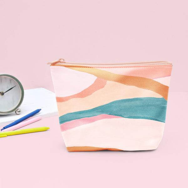 A hill-like design in pastels colors such as creams, pinks, and blues. Pouch has a peach colored zipper and is displayed with Jotter pens and a small clock in front of a light pink background.