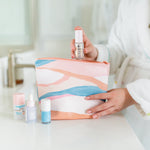 Lady in bathrobe placing self care products into a vegan leather light peach, blue, and pink pouch with a zip top.